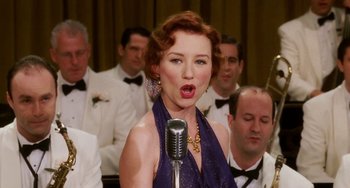 Movie still from “Mona Lisa Smile” (2003), directed by Mike Newell – A woman in a purple dress singing into a microphone in front of a group of men; Close Up shot, Low angle