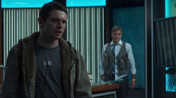 Movie still from “Money Monster” (2016), directed by Jodie Foster – A man standing next to another man holding a knife; Medium shot, Over the shoulder angle