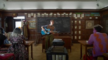 Movie still from “Monster High: The Movie” (2022), directed by Todd Holland – A man playing a guitar in front of a chalkboard; Wide shot, Over the shoulder angle