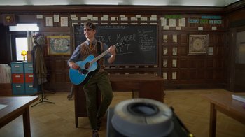 Movie still from “Monster High: The Movie” (2022), directed by Todd Holland – A man sitting in front of a chalkboard holding a guitar; Wide shot, Low angle