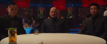 Movie still from “Mortdecai” (2015), directed by David Koepp – A man sitting at a table with a drink in front of him; Medium shot, Over the shoulder angle