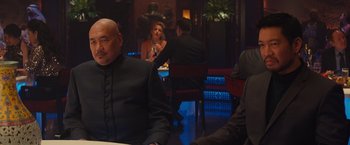 Movie still from “Mortdecai” (2015), directed by David Koepp – A group of people sitting at a table in a room; Medium shot, Over the shoulder angle