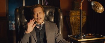 Movie still from “Mortdecai” (2015), directed by David Koepp – A man sitting in a chair waving at the camera; Medium shot, Over the shoulder angle