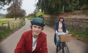 Movie still from “Mothering Sunday” (2021), directed by Eva Husson – Two women riding bicycles down a road wearing hats; Medium shot, Over the shoulder angle