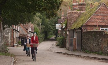 Movie still from “Mothering Sunday” (2021), directed by Eva Husson – Two women riding bikes down a street near a tree; Extreme Wide shot, Over the shoulder angle