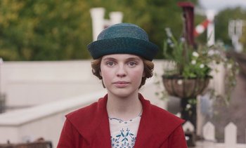 Movie still from “Mothering Sunday” (2021), directed by Eva Husson – A young woman wearing a green hat and a red jacket; Close Up shot, Low angle