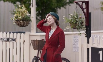 Movie still from “Mothering Sunday” (2021), directed by Eva Husson – A woman in a red coat standing next to a bicycle; Medium shot, Over the shoulder angle