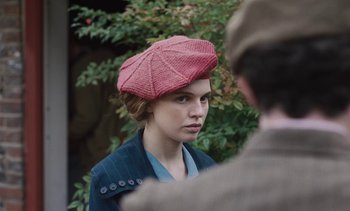 Movie still from “Mothering Sunday” (2021), directed by Eva Husson – A woman wearing a pink hat and a blue jacket; Close Up shot, Over the shoulder angle