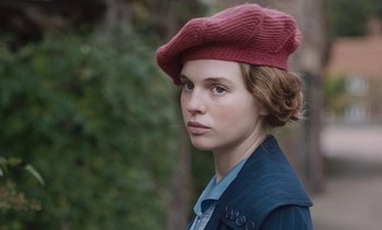Movie still from “Mothering Sunday” (2021), directed by Eva Husson – A young woman wearing a red hat and a blue jacket; Close Up shot, Over the shoulder angle