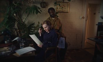 Movie still from “Mothering Sunday” (2021), directed by Eva Husson – A woman sitting at a table while a man stands next to her; Medium shot, High angle