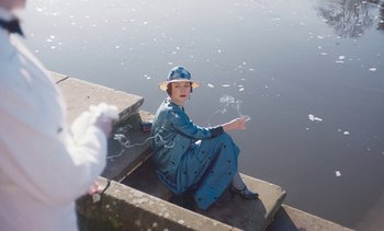 Movie still from “Mothering Sunday” (2021), directed by Eva Husson – An older woman sitting on the edge of the water; Medium shot, High angle