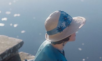 Movie still from “Mothering Sunday” (2021), directed by Eva Husson – A woman wearing a hat sitting on a pier; Close Up shot, High angle