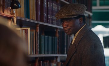 Movie still from “Mothering Sunday” (2021), directed by Eva Husson – A man wearing a hat standing in front of a book shelf; Close Up shot, Over the shoulder angle