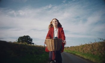 Movie still from “Mothering Sunday” (2021), directed by Eva Husson – A woman riding a bike down a road with trees in the background; Medium shot, Low angle