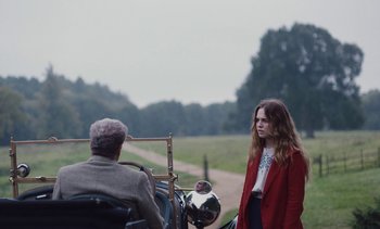 Movie still from “Mothering Sunday” (2021), directed by Eva Husson – A woman in a red coat standing next to a man in a wheelchair; Medium shot, Over the shoulder angle