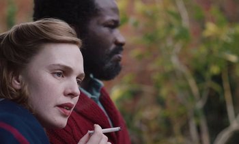 Movie still from “Mothering Sunday” (2021), directed by Eva Husson – A woman holding a cigarette in her mouth while standing next to a man; Close Up shot, Over the shoulder angle