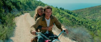 Movie still from “Mamma Mia! Here We Go Again” (2018), directed by Ol Parker – A man and a woman riding a motorcycle down a dirt road; Medium shot, High angle