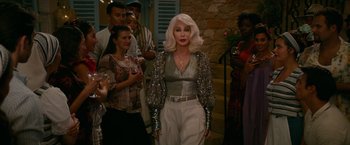 Movie still from “Mamma Mia! Here We Go Again” (2018), directed by Ol Parker – An older woman in a silver top is standing in front of a group of people; Medium shot, Over the shoulder angle