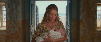 Movie still from “Mamma Mia! Here We Go Again” (2018), directed by Ol Parker – A woman holding a baby in her arms in a doorway; Medium shot, High angle
