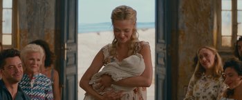 Movie still from “Mamma Mia! Here We Go Again” (2018), directed by Ol Parker – A woman holding a baby in her arms in front of a beach; Medium shot, Over the shoulder angle