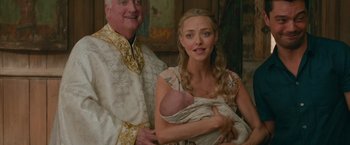 Movie still from “Mamma Mia! Here We Go Again” (2018), directed by Ol Parker – A man and woman holding a baby while wearing a priest's vestments; Close Up shot, High angle