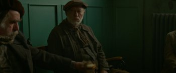 Movie still from “Mrs. Harris Goes to Paris” (2022), directed by Anthony Fabian – An older man sitting in a chair holding a sandwich; Medium shot, Over the shoulder angle