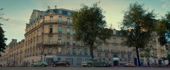 Movie still from “Mrs. Harris Goes to Paris” (2022), directed by Anthony Fabian – A tree in front of a building with cars parked in front of it; Extreme Wide shot, Low angle