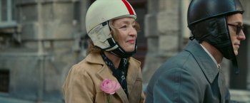 Movie still from “Mrs. Harris Goes to Paris” (2022), directed by Anthony Fabian – An older woman wearing a motorcycle helmet with a rose in it; Close Up shot, Over the shoulder angle