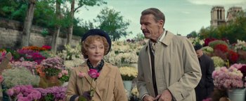 Movie still from “Mrs. Harris Goes to Paris” (2022), directed by Anthony Fabian – An older man standing next to an older woman holding a pink rose; Medium shot, Over the shoulder angle
