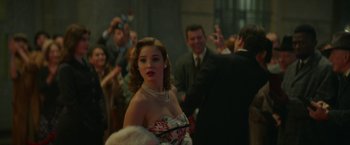 Movie still from “Mrs. Harris Goes to Paris” (2022), directed by Anthony Fabian – A beautiful woman in a dress and pearls in front of a group of people; Medium shot, Over the shoulder angle