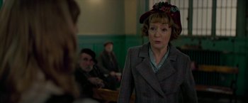 Movie still from “Mrs. Harris Goes to Paris” (2022), directed by Anthony Fabian – An older woman wearing a hat in front of a group of people; Medium shot, Over the shoulder angle