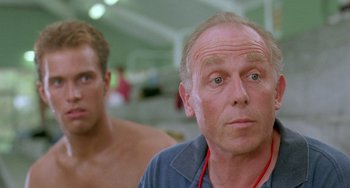 Movie still from “Muriel's Wedding” (1994), directed by P.J. Hogan – A man with a beard and a red necklace is next to another man with no shirt; Close Up shot, Low angle