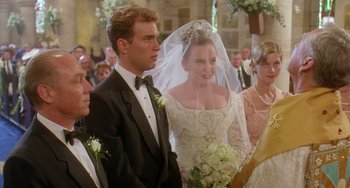 Movie still from “Muriel's Wedding” (1994), directed by P.J. Hogan – A man and woman in wedding attire standing next to each other in front of a group of people; Medium shot, Over the shoulder angle