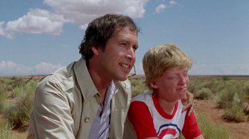 Movie still from “Vacation” (1983), directed by Harold Ramis – A man sitting next to a young boy in a field; Medium shot, Low angle