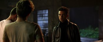 Movie still from “Need for Speed” (2014), directed by Scott Waugh – A man standing next to another man in a dark room; Medium shot, Over the shoulder angle