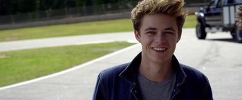Movie still from “Need for Speed” (2014), directed by Scott Waugh – A young man smiling for the camera while standing on the side of the road; Close Up shot, Over the shoulder angle