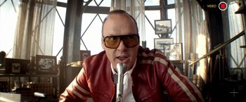 Movie still from “Need for Speed” (2014), directed by Scott Waugh – A man in a red jacket and sunglasses is sitting at a microphone; Close Up shot, Low angle