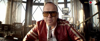Movie still from “Need for Speed” (2014), directed by Scott Waugh – A man in a red jacket and sunglasses is sitting at a microphone; Close Up shot, Low angle