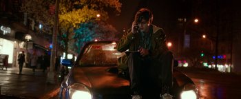 Movie still from “Nerve” (2016), directed by Henry Joost – A man sitting on the hood of a car while on a cell phone; Medium shot, Over the shoulder angle