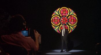 Movie still from “Network” (1976), directed by Sidney Lumet – A man in a suit and tie standing in front of a stained - glass window; Wide shot, Over the shoulder angle