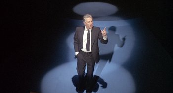 Movie still from “Network” (1976), directed by Sidney Lumet – A man wearing a suit and tie standing in a dark room; Wide shot, Low angle