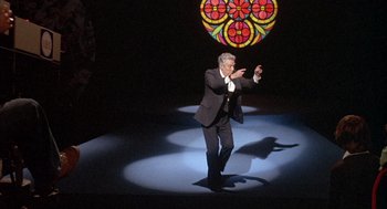 Movie still from “Network” (1976), directed by Sidney Lumet – A man in a black suit is holding a wand; Wide shot, High angle