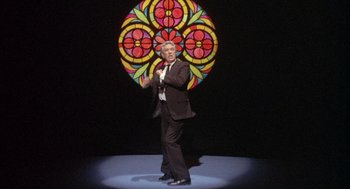 Movie still from “Network” (1976), directed by Sidney Lumet – A man standing on a stage in front of a stained - glass window; Wide shot, Low angle