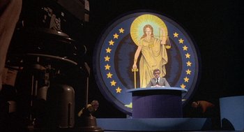 Movie still from “Network” (1976), directed by Sidney Lumet – A man is sitting at a podium in front of a statue of lady justice; Wide shot, Low angle