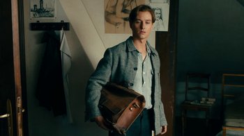 Movie still from “Never Look Away” (2018), directed by Florian Henckel von Donnersmarck – A man holding a brown leather bag in a room; Medium shot, Over the shoulder angle