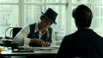 Movie still from “Never Look Away” (2018), directed by Florian Henckel von Donnersmarck – A man wearing a hat sitting in front of a window; Medium shot, Over the shoulder angle