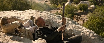 Movie still from “News of the World” (2020), directed by Paul Greengrass – An older man holding a rifle while sitting on top of a rock; Medium shot, High angle