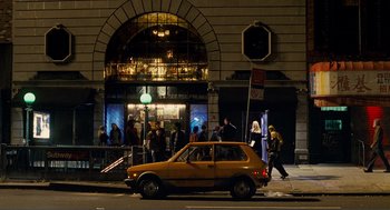 Movie still from “Nick and Norah's Infinite Playlist” (2008), directed by Peter Sollett – A car parked on the side of the road near a building; Wide shot, High angle