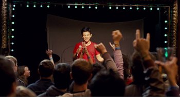 Movie still from “Nick and Norah's Infinite Playlist” (2008), directed by Peter Sollett – A man standing in front of an audience holding a microphone; Medium shot, Over the shoulder angle