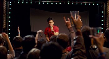 Movie still from “Nick and Norah's Infinite Playlist” (2008), directed by Peter Sollett – A man in a red t - shirt speaking to a crowd of onlookers; Medium shot, Over the shoulder angle